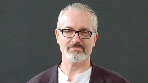 Man with grey hair, beard, and glasses smiling and wearing a white shirt and dark blazer.