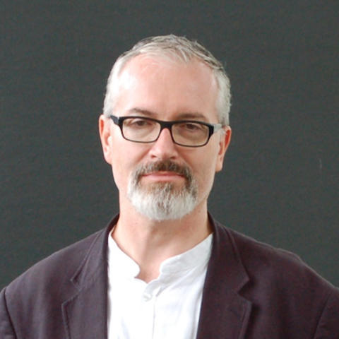 Man with grey hair, beard, and glasses smiling and wearing a white shirt and dark blazer.