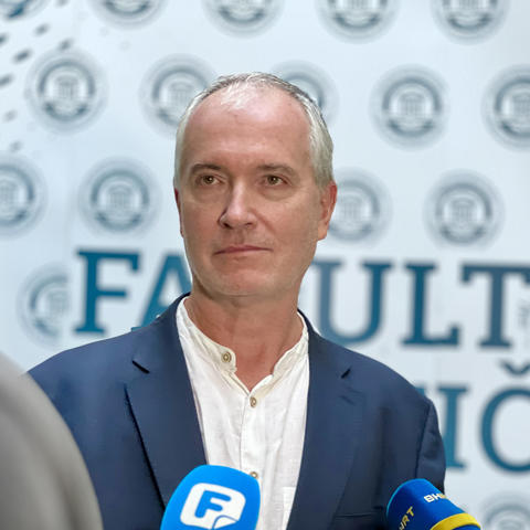 A man with grey hair, blue jacket, and white shirt is speaking into a news media microphone.