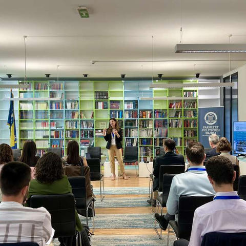 A woman stands in front of a large bookcase and speaks to an audience.