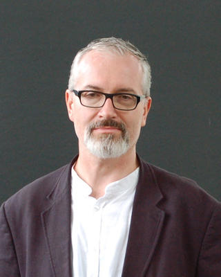 Man with grey hair, beard, and glasses smiling and wearing a white shirt and dark blazer.