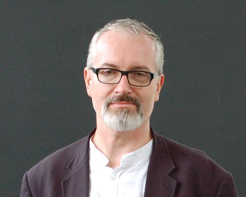 Man with grey hair, beard, and glasses smiling and wearing a white shirt and dark blazer.